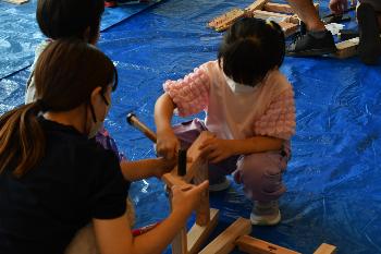 女の子とがお母さんに見守られながら金づちで釘を打ち込んでいる様子